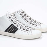 Three quarter front view of mid top white and black sneaker. Full leather upper with studs, an internal zip and vintage rubber sole.