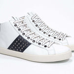 Three quarter front view of mid top white and navy sneaker. Full leather upper with studs, an internal zip and vintage rubber sole.