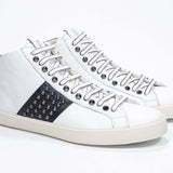 Three quarter front view of mid top white and navy sneaker. Full leather upper with studs, an internal zip and vintage rubber sole.