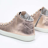 Three quarter back view of mid top rose-gold sneaker with full leather upper with perforated crown logo, internal zip and white sole.