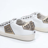 Three quarter back view of low top white and metallic gold sneaker. Full leather upper with studs and vintage rubber sole.