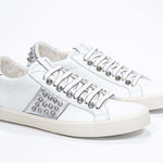 Three quarter front view of low top white and metallic silver sneaker. Full leather upper with studs and vintage rubber sole.