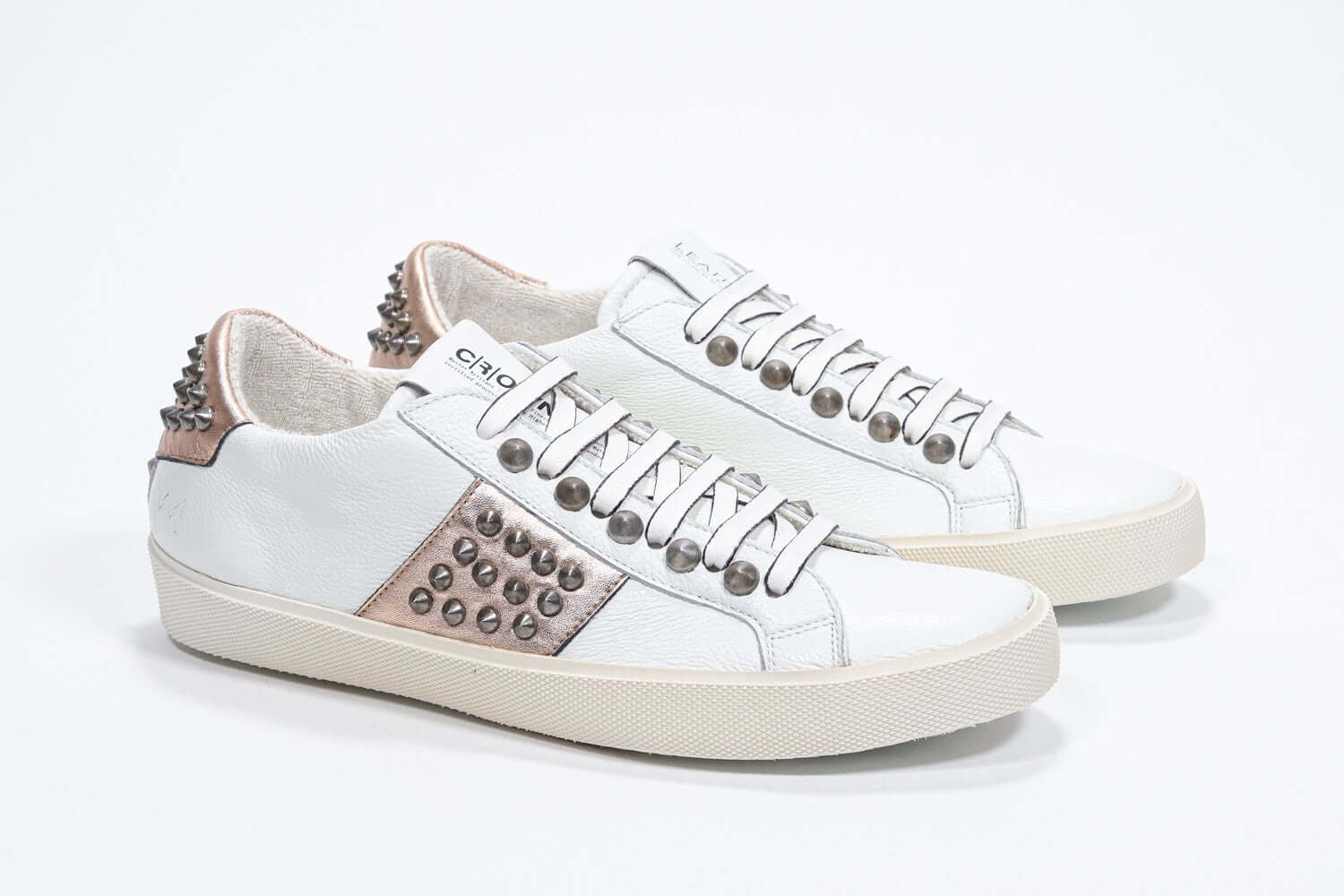 Three quarter front view of low top white and metallic rose sneaker. Full leather upper with studs and vintage rubber sole.