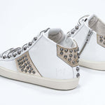 Three quarter back view of mid top white and metallic gold sneaker. Full leather upper with studs, an internal zip and vintage rubber sole.
