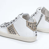 Three quarter back view of mid top white and metallic gold sneaker. Full leather upper with studs, an internal zip and vintage rubber sole.